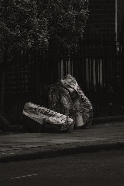 A pile of large, black rubbish bags made from thick plastic, filled with waste materials, are stacked on the pavement beside a dark metal fence with vertical bars. The bags appear slightly wrinkled an