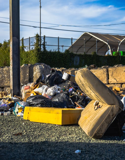 A pile of large, black rubbish bags made from thick plastic, filled with waste materials, are stacked on the pavement beside a dark metal fence with vertical bars. The bags appear slightly wrinkled an