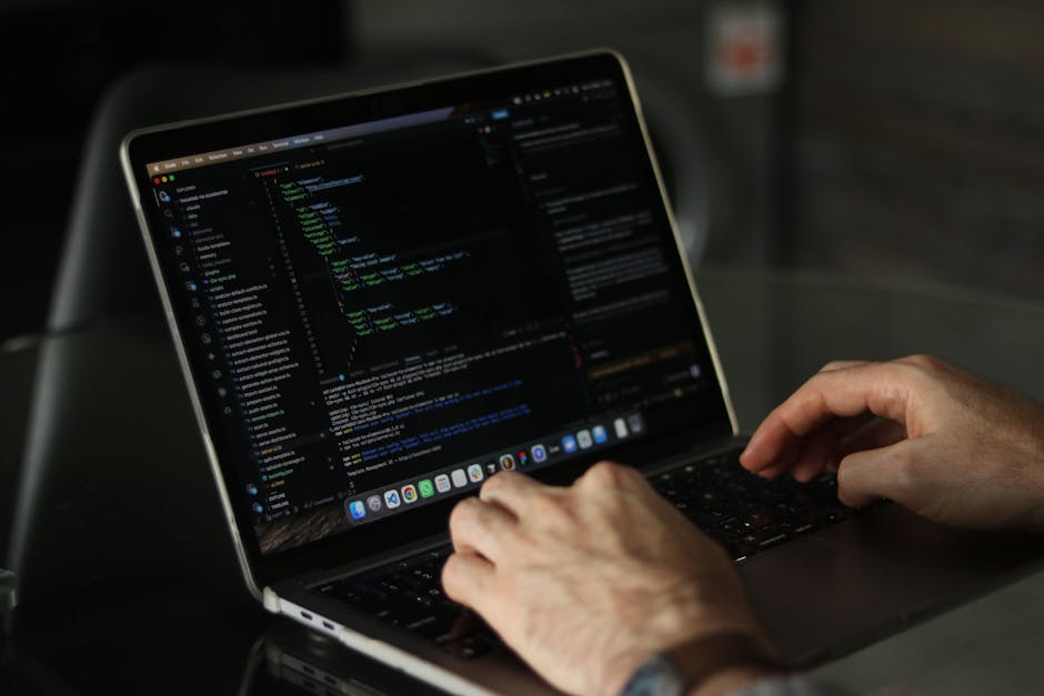 A pair of adult hands with light skin and visible veins are positioned over a laptop keyboard, actively typing. The laptop, with a silver or metallic finish, is open on a dark table or desk surface. T
